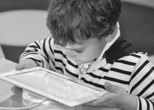 Photo of a young boy holding a computer tablet close to his face sitting at a desk