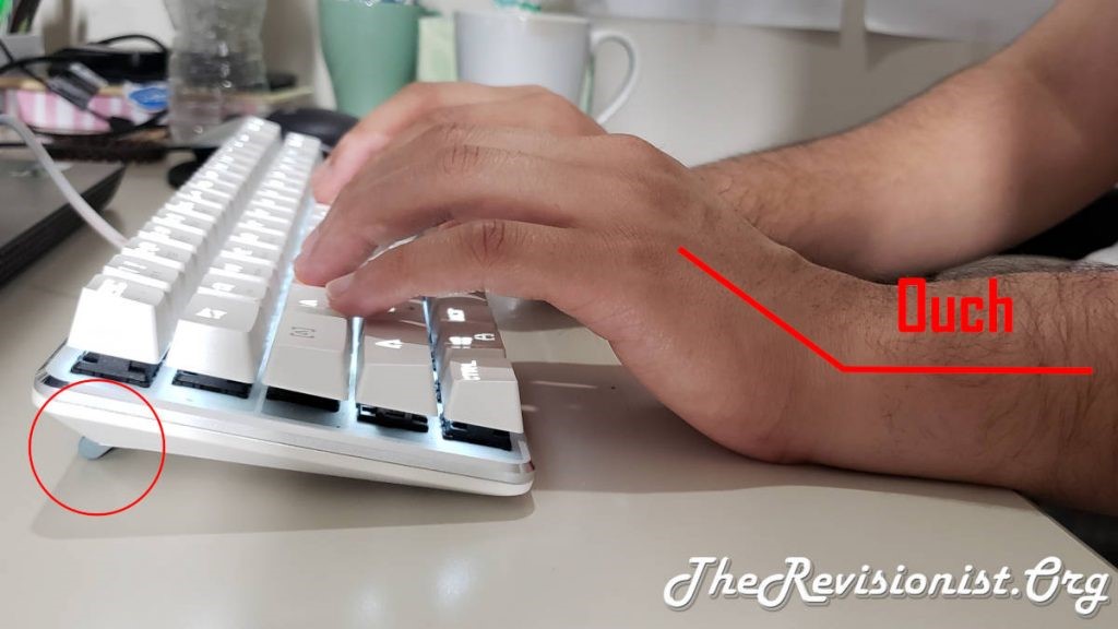 Photo showing a keyboard with feet up in the back and a person's hands bent up to use it