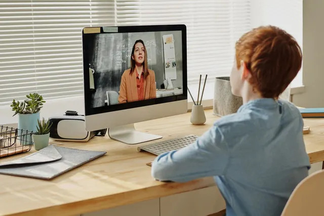 Photo of a student at a table looking at a monitor showing a video of their teacher